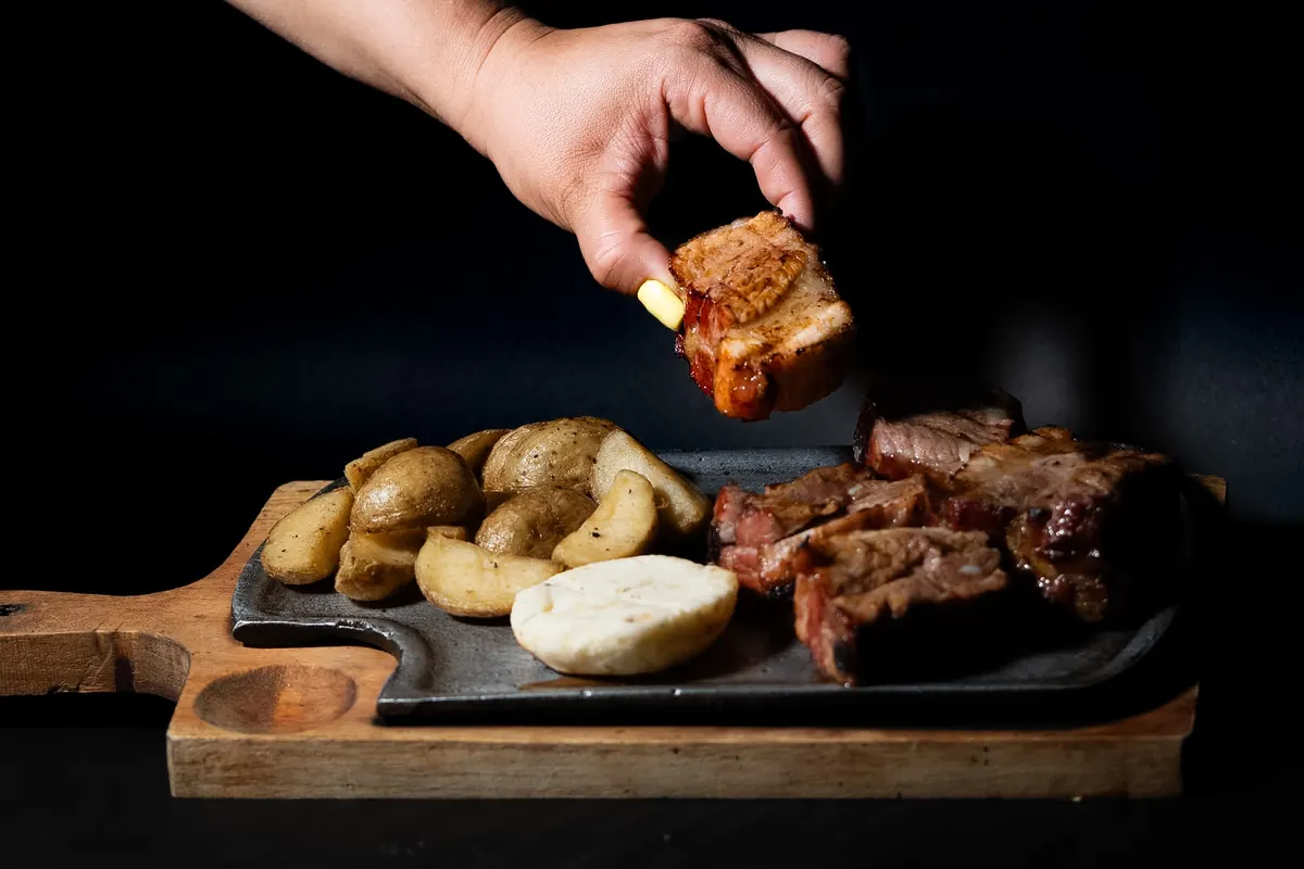 Mano tomando un trozo de costilla al barril de una tabla con papas en cascos y arepa